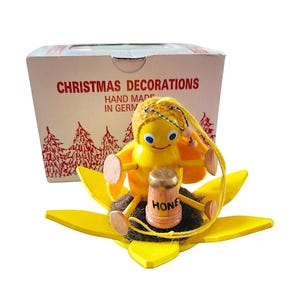 May include: A yellow wooden bee ornament with blue eyes and a textured head, sitting on a yellow flower base. The bee holds a small wooden honey pot labeled HONEY. Behind the ornament is a white box labeled CHRISTMAS DECORATIONS HAND MADE IN GERMANY.
