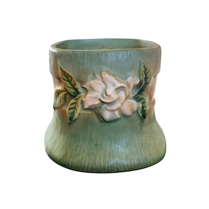 May include: A ceramic planter featuring a muted sage green base with a vertical ribbed texture. The exterior is adorned with a raised, three-dimensional relief of a pale pink flower surrounded by green leaves, creating a decorative botanical accent.