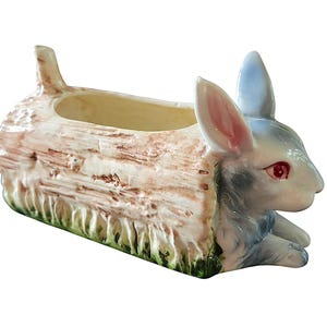 May include: A ceramic planter shaped like a hollow log with a textured bark finish in shades of tan and brown. A rabbit with blue-toned fur, pink-lined ears, and red eyes is attached to the front, with green grass detailing at the base.