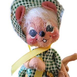 Może przedstawiać: Zabytkowa pluszowa lalka myszy z malowaną twarzą, w zielono-białej kraciastej czapeczce i sukience z akcentami słonecznik&oacute;w. Lalka ma r&oacute;żowe uszy, białe włosy i ż&oacute;łtą wstążkę.
