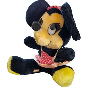 May include: Vintage plush toy featuring a cartoon character with a yellow face, black ears, and a black nose. The toy wears a pink polka-dot dress and a red bow. The feet are yellow.