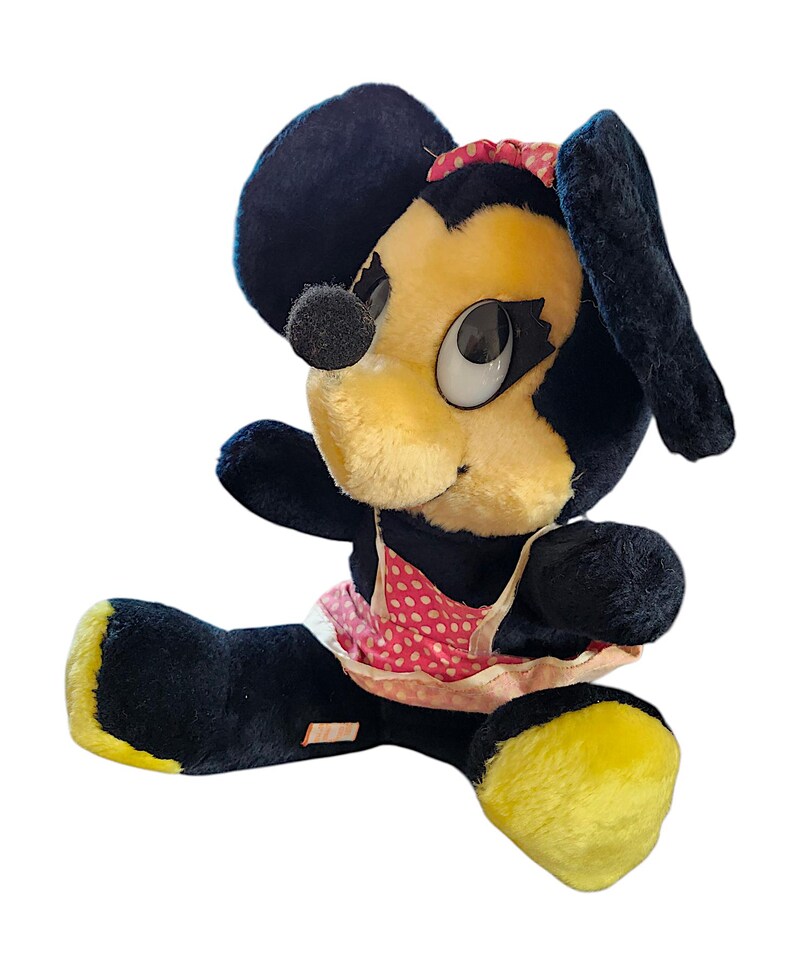 May include: Vintage plush toy featuring a cartoon character with a yellow face, black ears, and a black nose. The toy wears a pink polka-dot dress and a red bow. The feet are yellow.