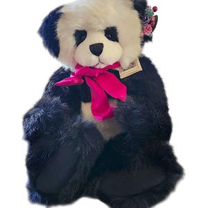 May include: A plush panda bear with black and white fur, featuring a bright pink satin ribbon tied around its neck. The bear has small fabric flowers attached near its right ear and a small rectangular paper tag attached to its chest area.