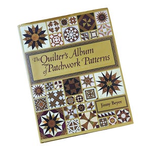 May include: A hardcover book titled The Quilter's Album of Patchwork Patterns by Jinny Beyer. The cover features a gold background with a collage of various geometric quilt block designs in shades of brown, purple, and white throughout.