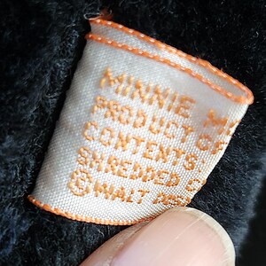 May include: Close-up of a white tag with orange trim and text that reads "MINNIE M PRODUCT OF CONTENTS SHREDDED @ WALT DISNEY". The tag is partially obscured by a dark, textured fabric.