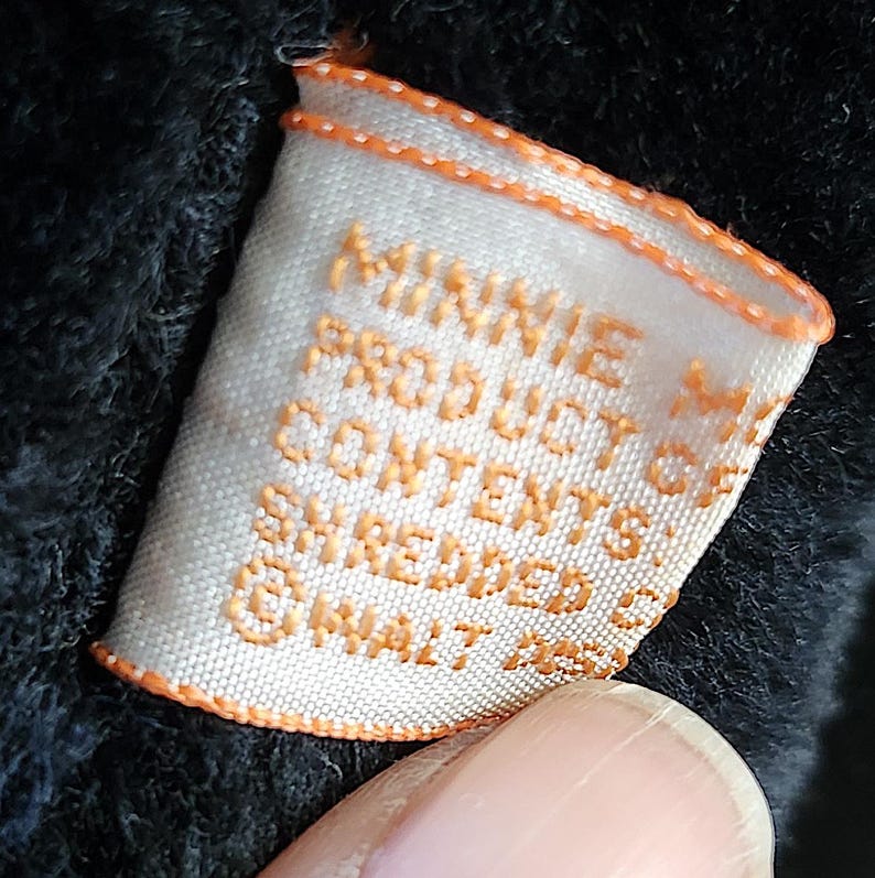 May include: Close-up of a white tag with orange trim and text that reads "MINNIE M PRODUCT OF CONTENTS SHREDDED @ WALT DISNEY". The tag is partially obscured by a dark, textured fabric.