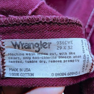 Puede incluir: Una prenda de tela color granate con una etiqueta de cuidado blanca en el interior que muestra el logotipo de Wrangler, el n&uacute;mero de modelo 936CWE y la talla 29 x 32. La etiqueta incluye instrucciones de lavado, Made in USA, 100% algod&oacute;n y c&oacute;digo.