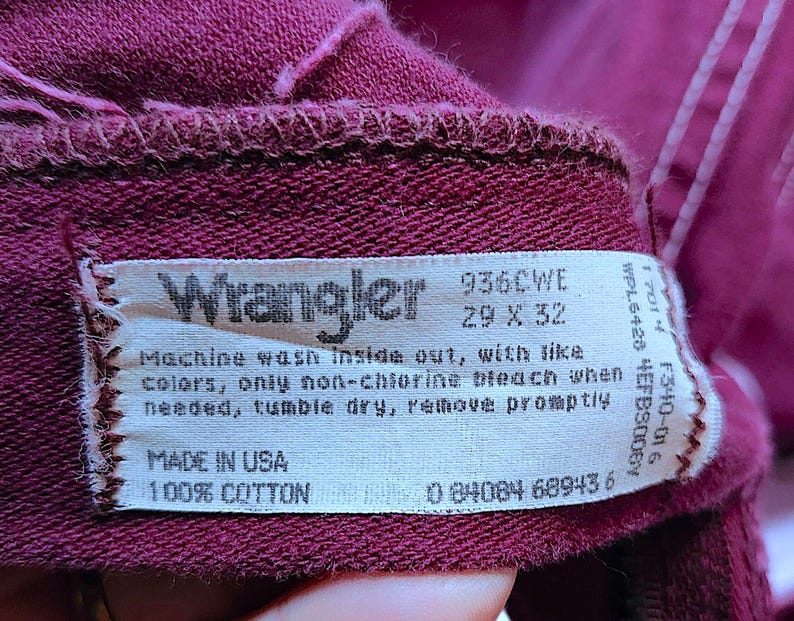 Puede incluir: Una prenda de tela color granate con una etiqueta de cuidado blanca en el interior que muestra el logotipo de Wrangler, el n&uacute;mero de modelo 936CWE y la talla 29 x 32. La etiqueta incluye instrucciones de lavado, Made in USA, 100% algod&oacute;n y c&oacute;digo.