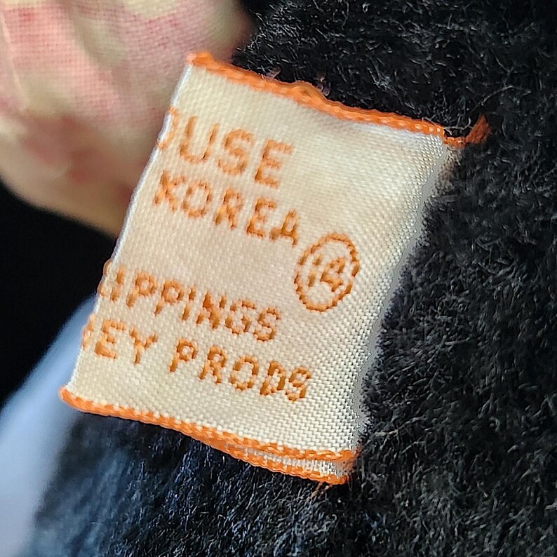 May include: Close-up of a rectangular tag with orange trim and text. The tag reads "USE KOREA" and "LIPPINGS EY PRODS". The tag is attached to a black, fuzzy material. A small, circular symbol with the number 14 is also visible.