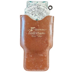 May include: A vintage Jeppesen Slide Graphic Computer in a brown leather case. The slide rule is white with black and green markings. The case has the words "Jeppesen Slide Graphic Computer" embossed on it. The case is brown with a worn appearance.