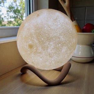 May include: A spherical, illuminated moon lamp with a textured surface, resting on a brown, curved stand. The lamp emits a soft, warm glow, creating a celestial ambiance. The lamp is on a wooden surface.