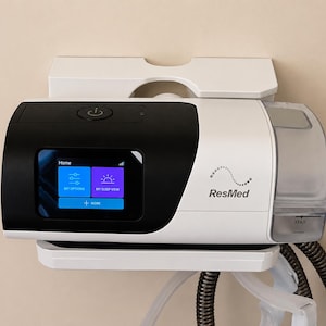 May include: A white and black CPAP machine with a digital display showing menu options. The machine is mounted on a white wall bracket. The brand name "ResMed" is visible on the side.