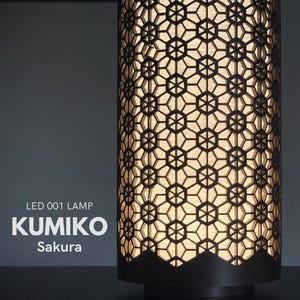 Op de afbeelding: Cilindrische lamp met een geometrisch patroon. De lamp heeft een donkerbruine buitenkant met een complex, opengewerkt ontwerp waardoor licht doorheen schijnt. De tekst "KUMIKO Sakura" is zichtbaar op de zijkant van de lamp.