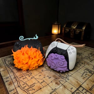 May include: Two video game controllers customized with decorative attachments. One black controller features a bright orange flame-like design and a light blue swirl. The other white controller has a purple cloud-like attachment and a tan curved stem.