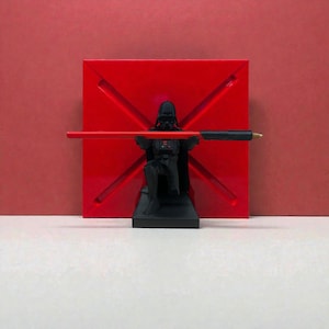 Puede incluir: Un portalápices negro de Darth Vader con un bolígrafo rojo, sobre un fondo cuadrado rojo. El portalápices es una figura detallada de Darth Vader. El fondo es un cuadrado rojo liso con un diseño en X.