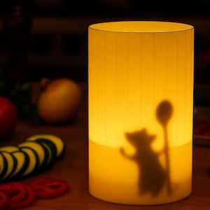 Pode incluir: Lâmpada cilíndrica iluminada com um brilho amarelo quente. A lâmpada apresenta uma silhueta de um rato segurando uma colher. Vegetais fatiados e outros itens de cozinha são visíveis no fundo.