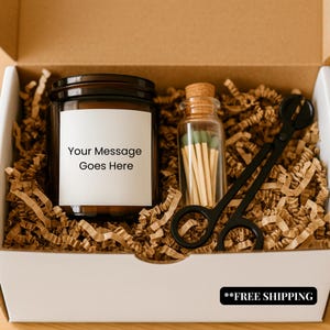 May include: A gift box containing a brown glass jar candle with a white label that reads "Your Message Goes Here", a small glass jar of matches, and black candle wick trimmers. The box is filled with brown crinkle paper.