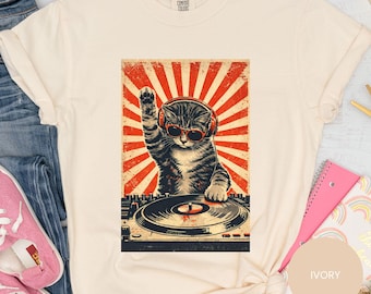 May include: Ivory-colored t-shirt featuring a graphic of a cat DJ wearing headphones and sunglasses, with a red and white sunburst background. The cat is raising a paw. The shirt has a relaxed fit.