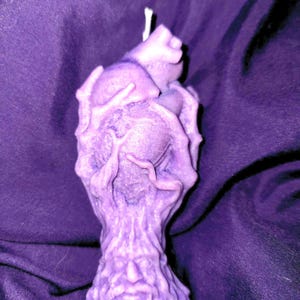 May include: A purple heart-shaped candle with a detailed, textured design resembling a hand holding the heart. The candle has a white wick.