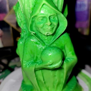 May include: Green candle shaped like a person wearing a hooded robe and holding a sphere. The candle is on a white base.