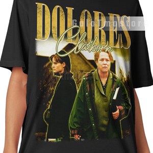 Puede incluir: Camiseta negra con el título de la película "Dolores Claiborne" en letras doradas. La camiseta muestra una imagen vintage de dos mujeres. El texto "Dolores Claiborne" está en una fuente estilizada.