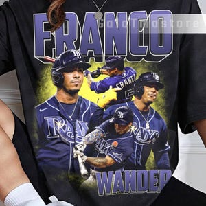 Peut inclure: T-shirt noir avec un collage du joueur de baseball Wander Franco. Le design comprend le nom "FRANCO" en grandes lettres violettes et le nom "WANDER" en violet en dessous des images. Le t-shirt a un style vintage.