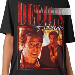 May include: Black t-shirt featuring a graphic design with the text "DEVILS Advocate" in red and white. The design includes two images of actors. The shirt is a unisex style.