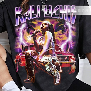 May include: Black graphic t-shirt featuring the text "KALI UCHIS" in a purple, retro font. The design includes an illustration of a woman holding a microphone, a red car, palm trees, and lightning bolts. The shirt is unisex.