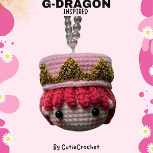 May include: A pink and gold crocheted doll with a crown, inspired by G-Dragon. The doll is hanging from a string of pearls. The text "G-DRAGON INSPIRED" is written above the doll. The text "By CutieCrochet" is written below the doll.