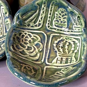 May include: A handmade ceramic dish with a four-sectioned design. Each section features a different embossed design, including a flower, crown, and geometric patterns. The dish is glazed in shades of green and blue.