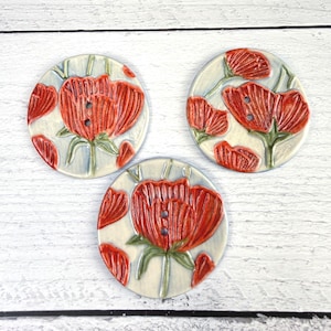May include: Three round ceramic buttons featuring a hand-painted design of vibrant orange-red poppy flowers with green stems and leaves against a pale, off-white background. Each button has two central holes for attachment and a smooth finish.