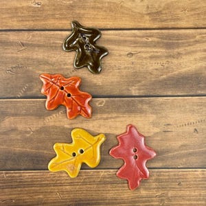 May include: Four ceramic buttons shaped like oak leaves in autumn colors. One button is dark brown, one is orange, one is yellow, and one is red. Each button features two holes in the center for attachment and a glossy, smooth finish on the surface.