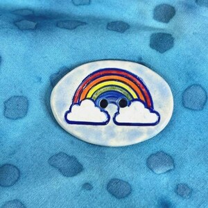 May include: A ceramic button with a rainbow and clouds design. The button is oval shaped and has a light blue background with white clouds and a rainbow with red, orange, yellow, green, blue, indigo, and violet colors.
