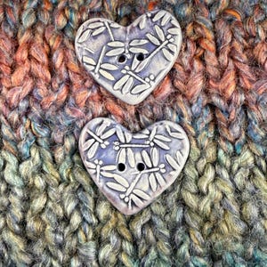 May include: Two heart-shaped ceramic buttons with dragonfly designs. The buttons are light purple with white dragonfly details and black buttonholes. They are set against a backdrop of colorful, textured yarn in shades of orange, blue, and green.