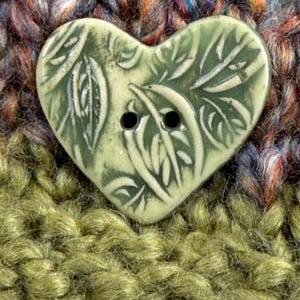May include: A heart-shaped button with a light green glaze and a leaf design. The button has two black holes for sewing and is set against a textured, multicolored background of yarn. The button is a craft supply.