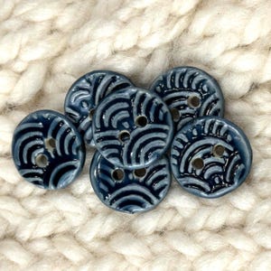 May include: A collection of six round, dark blue ceramic buttons, each featuring a glossy finish and a raised, light blue wave-like pattern. Every button is designed with two central holes for attachment, showcasing a consistent circular shape.
