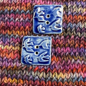 May include: Two square, cobalt blue ceramic buttons with a raised bird design and three holes. The buttons are set against a backdrop of colorful, hand-knitted fabric in shades of purple, pink, orange, and brown.