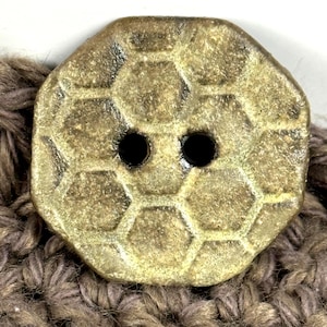 May include: A close-up of a decorative button with a hexagonal pattern and two black holes. The button is a muted gold color and is attached to a brown knitted fabric. The button is octagonal in shape.