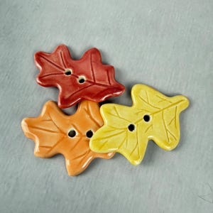May include: Three leaf-shaped buttons in fall colors: red, orange, and yellow. Each button has two holes for sewing. The buttons are made of a glossy material and are arranged on a light gray surface. These decorative buttons are perfect for autumn-themed crafts.