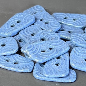 May include: A pile of light blue ceramic buttons with a textured, wave-like pattern. Each button has two holes for sewing. The buttons are irregularly shaped, with rounded edges and a slightly speckled appearance.