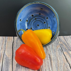May include: A round, blue ceramic colander with drainage holes, containing a yellow bell pepper. A red bell pepper and an orange bell pepper are positioned in front of the colander. The ceramic bowl features a mottled blue glaze finish.