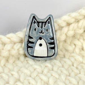 May include: A ceramic button in the shape of a cat. The button is light blue with black stripes and details. The cat has a white belly and a small black button. The button is set against a cream-colored, textured background.