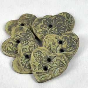 May include: A collection of heart-shaped buttons with a distressed, antique appearance. The buttons are primarily olive green with darker, detailed floral patterns. Each button has two holes for sewing.