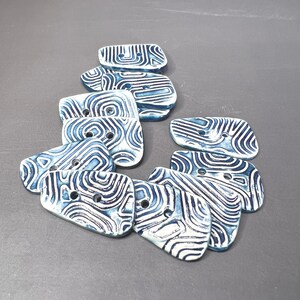 May include: A collection of ceramic buttons in various sizes, featuring a blue and white swirled pattern. Each button has two holes for sewing. The buttons are arranged on a gray surface, showcasing their unique design and texture.