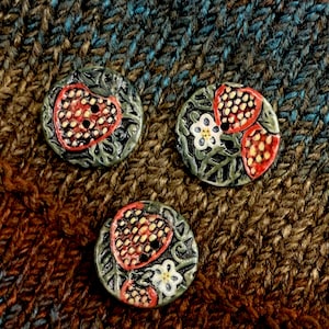 May include: Three ceramic buttons with a strawberry design. Each button features a red strawberry with black seeds, a white flower, and green leaves. The buttons are round and have two small black holes for sewing.