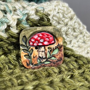 May include: A ceramic button with a hand-painted design of a red and white mushroom with green leaves on a brown background.