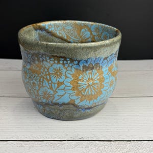 May include: A ceramic cup featuring a light blue floral pattern set against a textured, earthy brown background. The rim and base are finished in a muted, mottled grey-brown glaze, with the floral design wrapping around the exterior surface.