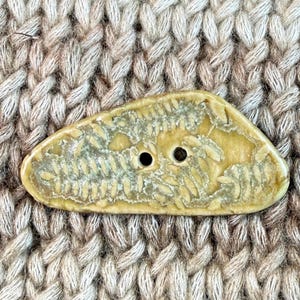 May include: A light yellow, oval-shaped button with two holes. The button has a textured surface with a leaf-like pattern and is set against a background of knitted fabric in shades of beige and brown.