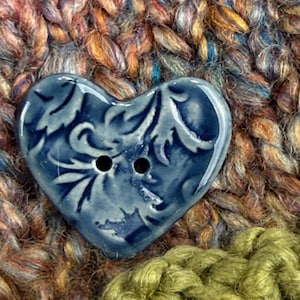 May include: A blue heart-shaped button with a floral design and two holes. The button is set against a backdrop of textured, multicolored yarn in shades of brown, orange, and green, creating a cozy, handcrafted aesthetic.