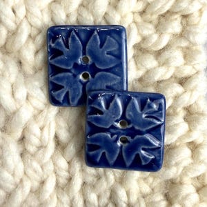 May include: Two square, cobalt blue ceramic buttons with a raised floral design. Each button has two holes for sewing. The buttons are set against a background of thick, off-white knitted fabric.